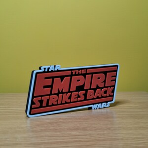 May include: A black and white 3D printed sign with the text "STAR WARS THE EMPIRE STRIKES BACK" in red letters.