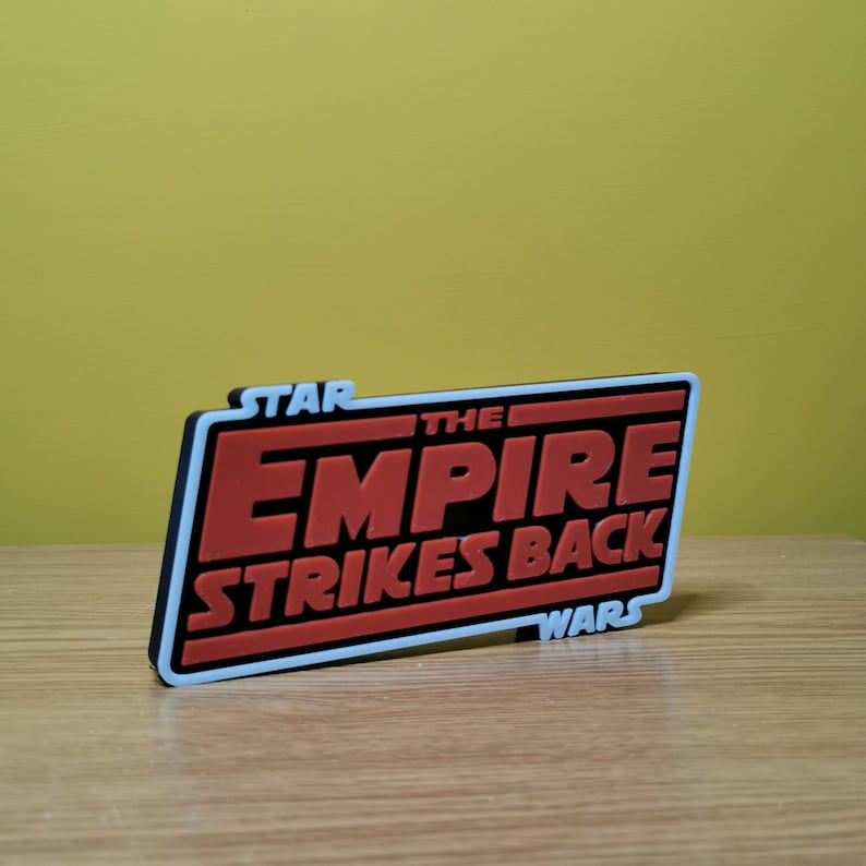 May include: A black and white 3D printed sign with the text "STAR WARS THE EMPIRE STRIKES BACK" in red letters.
