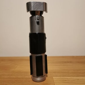K&ouml;nnte beinhalten: Ein silberner und schwarzer Lichtschwert-Requisit mit einem strukturierten Griff und einem schwarzen Griff. Das Lichtschwert hat eine silberne Kappe oben und eine silberne Basis.