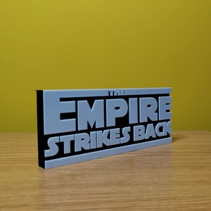 May include: A gray 3D printed sign with black lettering that reads "THE EMPIRE STRIKES BACK".