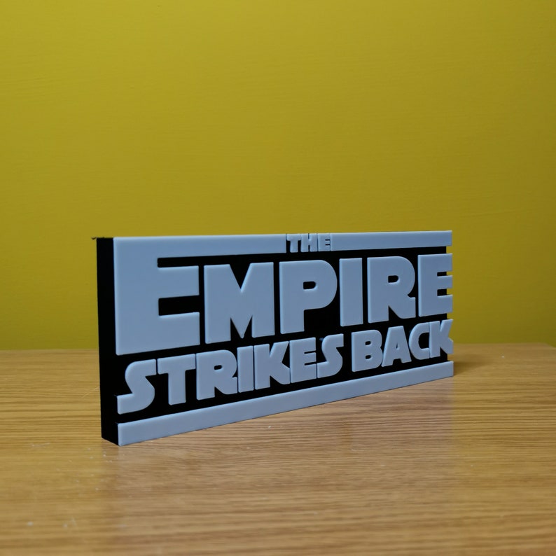 May include: A gray 3D printed sign with black lettering that reads "THE EMPIRE STRIKES BACK".