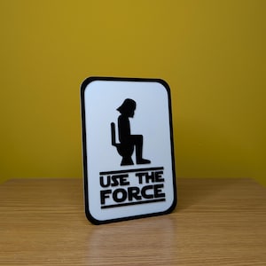 Puede incluir: Un cartel blanco con una silueta negra de Darth Vader sentado en un inodoro. El cartel dice "Use the Force" en letras negras.