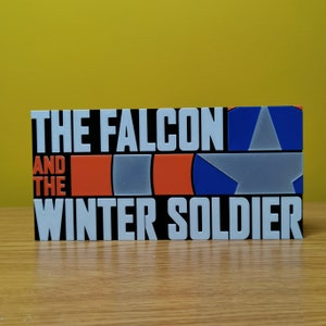 May include: A black and white sign with the text "The Falcon and the Winter Soldier" in white letters. The sign also features a red, white, and blue design with a star.