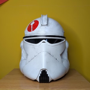 May include: A white helmet with a red emblem on top, featuring a black visor and a detailed, textured surface. The helmet has a distinctive design with a black trim and a black lower section.