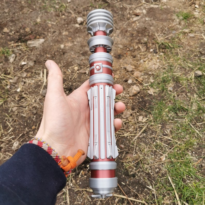 May include: A silver and red lightsaber hilt with a textured design. The hilt has a black button and a silver cap on the end.