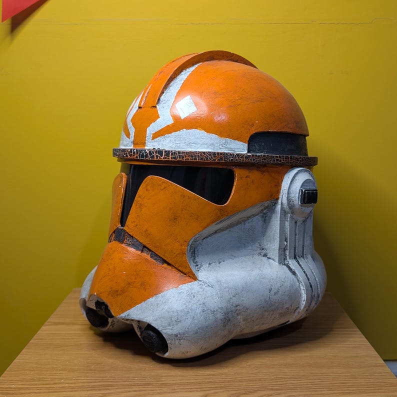 May include: An orange and white plastic clone trooper helmet with a weathered finish. The helmet has a white symbol on the top and a black visor.