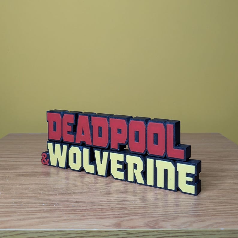 Może przedstawiać: Tr&oacute;jwymiarowa tabliczka z napisem "DEADPOOL & WOLVERINE" w czarnych, czerwonych i ż&oacute;łtych literach.