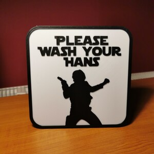 K&ouml;nnte beinhalten: Ein wei&szlig;es Schild mit schwarzem Rand und schwarzem Text, der "Please Wash Your Hans" lautet, mit einer Silhouette von Han Solo aus Star Wars, der einen Blaster h&auml;lt.