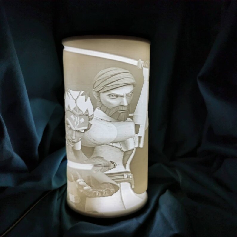 May include: A white ceramic lamp with a 3D image of two characters from the Star Wars: The Clone Wars animated series. The characters are a Jedi Master and his Padawan, both holding lightsabers.