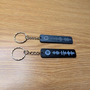 May include: Two black keychains with silver rings and chains. Each keychain has a white Spotify logo and a white barcode printed on the black rectangular tag.