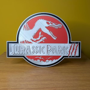 Op de afbeelding: Zilver en rood Jurassic Park logo, met een dinosaurus silhouet en de tekst "JURASSIC PARK". Het logo staat tegen een rode achtergrond met een zwarte rand en ligt op een houten oppervlak.