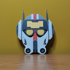 Pu&ograve; includere: Un casco stampato in 3D bianco, grigio e rosso con una visiera gialla. Il casco ha un design che ricorda un casco da pilota.
