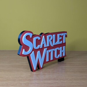May include: A 3D-printed sign with the words "SCARLET WITCH" in white, outlined in red, and with a black border. The sign is angled slightly, showcasing its layered design. The background is a solid yellow.