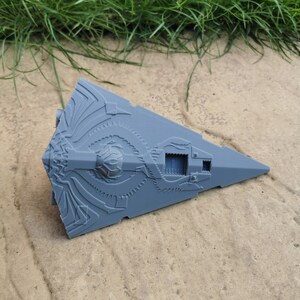 May include: A gray, detailed model of a spaceship with intricate designs and a small window on the side. The model is lying on a sandy surface.