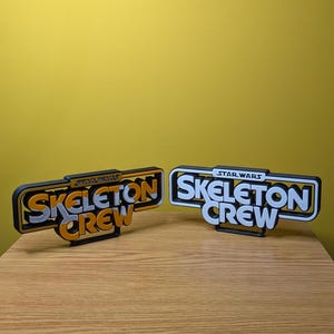 May include: Two black and white 3D printed signs with the text "Skeleton Crew" in orange and white letters. The top sign has the text "Star Wars" in small white letters above the text "Skeleton Crew". The bottom sign has the text "Star Wars" in small white letters above the text "Skeleton Crew".