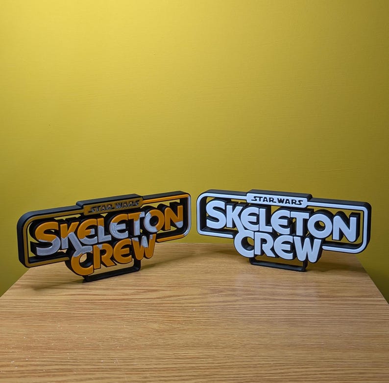 May include: Two black and white 3D printed signs with the text "Skeleton Crew" in orange and white letters. The top sign has the text "Star Wars" in small white letters above the text "Skeleton Crew". The bottom sign has the text "Star Wars" in small white letters above the text "Skeleton Crew".