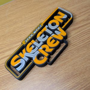 May include: A 3D printed sign with the words "Skeleton Crew" in a bold, stylized font. The words are in orange and silver, with a black outline. The sign also includes the words "Star Wars" in smaller text.