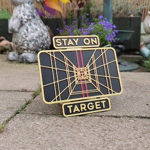Può includere: Un cartello in legno nero e dorato con una grafica di bersaglio a tema Star Wars. Il cartello recita "Rimani sul bersaglio".