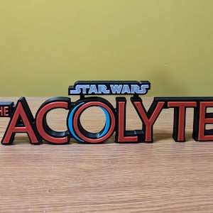 May include: A 3D printed logo for the Star Wars series "The Acolyte". The logo is black with red lettering and a blue and red circle in the center.