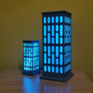 May include: Two modern, geometric lamps with a skyscraper design. The lamps are made of a dark gray material and have a blue light shining through the windows. The larger lamp is taller than the smaller lamp.