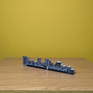 Peut inclure: Logo "WandaVision" noir et argent, probablement un objet d&eacute;coratif ou de collection. Le logo est en 3D, avec un design en couches et une police stylis&eacute;e. Il est pos&eacute; sur une surface en bois sur fond jaune.