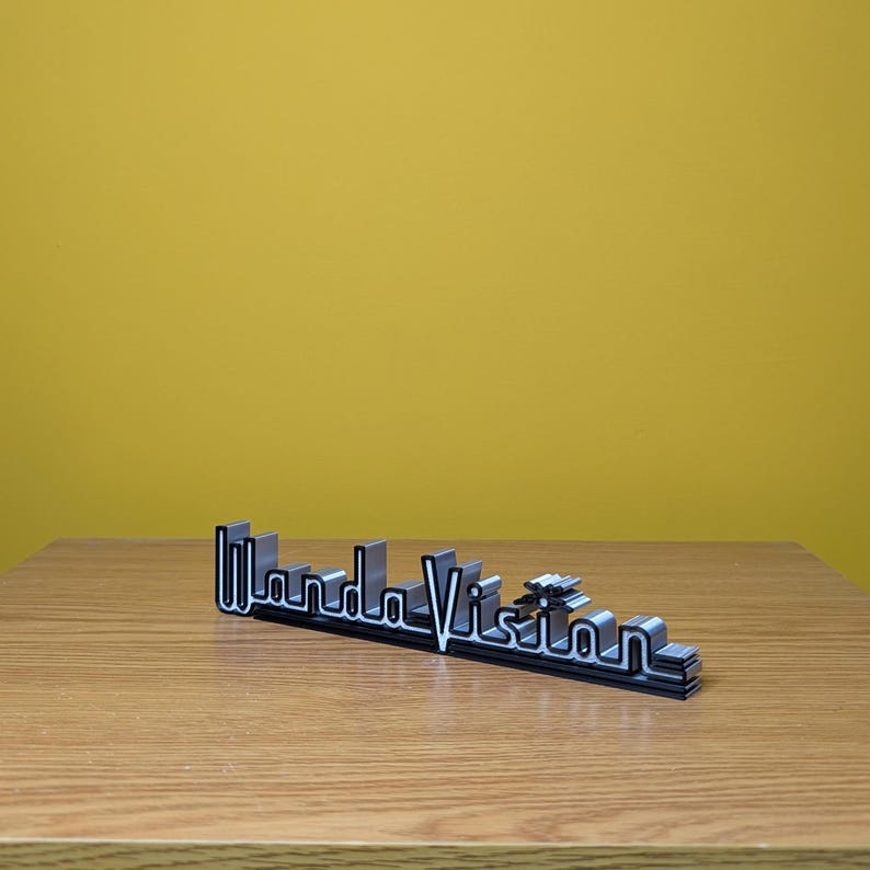 Peut inclure: Logo "WandaVision" noir et argent, probablement un objet d&eacute;coratif ou de collection. Le logo est en 3D, avec un design en couches et une police stylis&eacute;e. Il est pos&eacute; sur une surface en bois sur fond jaune.