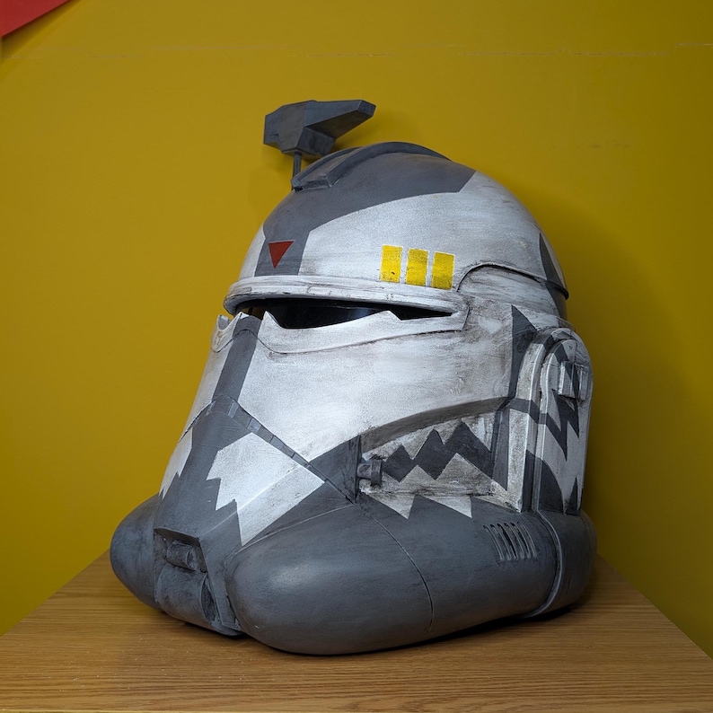 May include: A gray and white helmet with a black and white pattern. The helmet has a yellow stripe and a red triangle on the front. The helmet is made of plastic and is a replica of a Star Wars character.