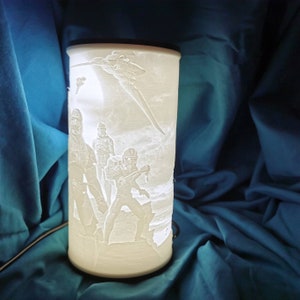 May include: A white ceramic lamp with a 3D design of Star Wars characters. The lamp features a scene of four characters standing in front of a spaceship. The characters are in a snowy landscape.