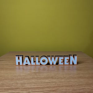 K&ouml;nnte beinhalten: Ein wei&szlig;es "HALLOWEEN"-Schild mit schwarzen Umrandungen und orangefarbenen Akzenten auf einer Holzoberfl&auml;che.