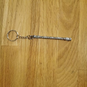 Op de afbeelding: Een zilveren metalen sleutelhanger met een gedetailleerd ontwerp dat lijkt op een futuristisch wapen. De sleutelhanger heeft een zilveren ring en een zilveren ketting.