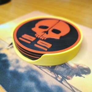 Puede incluir: Un juego de cuatro posavasos negros y naranjas con un dise&ntilde;o de calavera y huesos cruzados. Los posavasos est&aacute;n apilados y tienen una base amarilla.