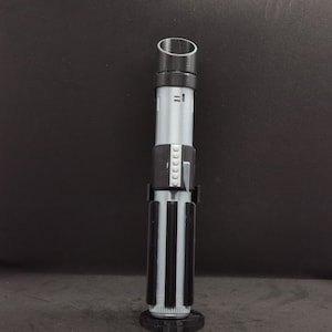 May include: A black and silver lightsaber toy with a black handle and a silver blade. The handle has a black ring with a series of small gray circles.