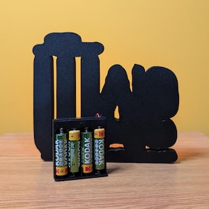 May include: Black silhouette of a person with the word "Love" in front of them. The silhouette is made of wood and is sitting on a wooden surface. There are four batteries in a battery pack in front of the silhouette.