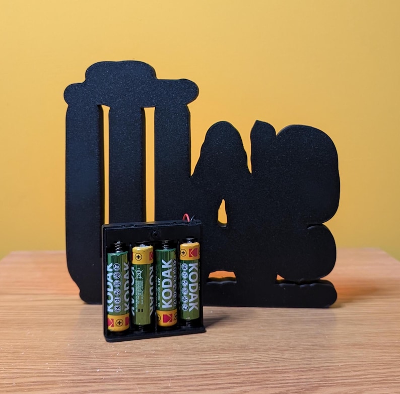 May include: Black silhouette of a person with the word "Love" in front of them. The silhouette is made of wood and is sitting on a wooden surface. There are four batteries in a battery pack in front of the silhouette.