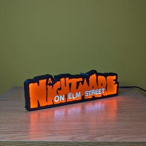 May include: A black and orange LED sign with the text "A NIGHTMARE ON ELM STREET" in a stylized font. The sign is illuminated, casting an orange glow. The sign is on a wooden surface.