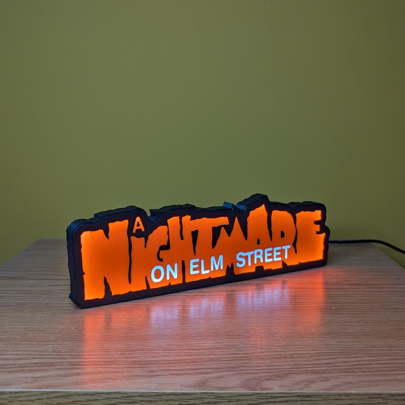 May include: A black and orange LED sign with the text "A NIGHTMARE ON ELM STREET" in a stylized font. The sign is illuminated, casting an orange glow. The sign is on a wooden surface.