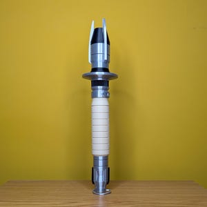 May include: A silver and white lightsaber prop with a black handle and a silver blade. The lightsaber is standing upright on a wooden surface.
