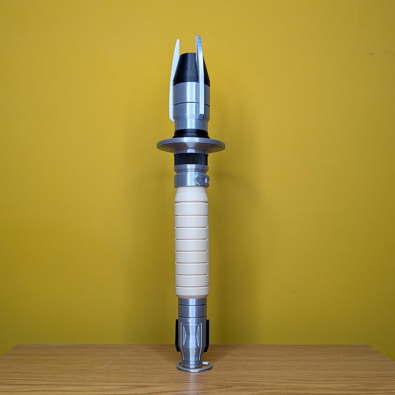 May include: A silver and white lightsaber prop with a black handle and a silver blade. The lightsaber is standing upright on a wooden surface.