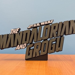 Op de afbeelding: Een zwart en grijs Star Wars-bord met de tekst "The Mandalorian and Grogu". Het 3D-bord staat op een zwarte basis. De woorden "Star Wars" staan bovenaan het bord.