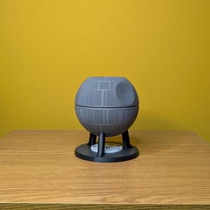 May include: A gray 3D printed model of the Death Star from Star Wars. The model is on a wooden surface.