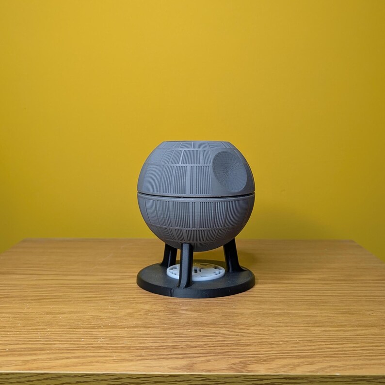 May include: A gray 3D printed model of the Death Star from Star Wars. The model is on a wooden surface.