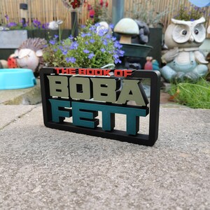 Peut inclure: Un panneau noir encadr&eacute; avec le texte "THE BOOK OF BOBA FETT" en lettres vertes, rouges et blanches. Le panneau est pos&eacute; sur une surface en b&eacute;ton gris.
