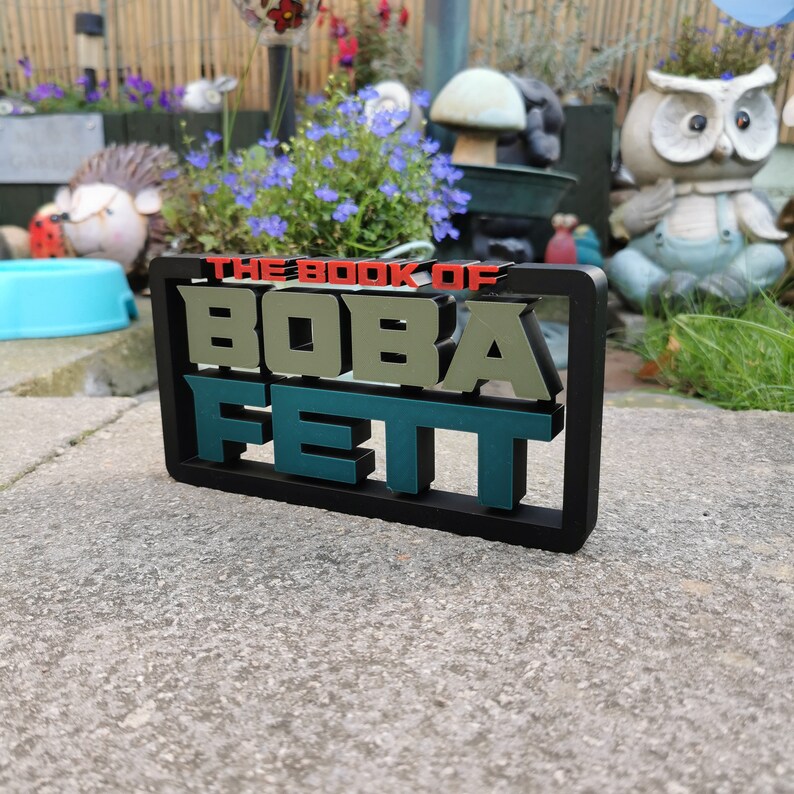 Peut inclure: Un panneau noir encadr&eacute; avec le texte "THE BOOK OF BOBA FETT" en lettres vertes, rouges et blanches. Le panneau est pos&eacute; sur une surface en b&eacute;ton gris.
