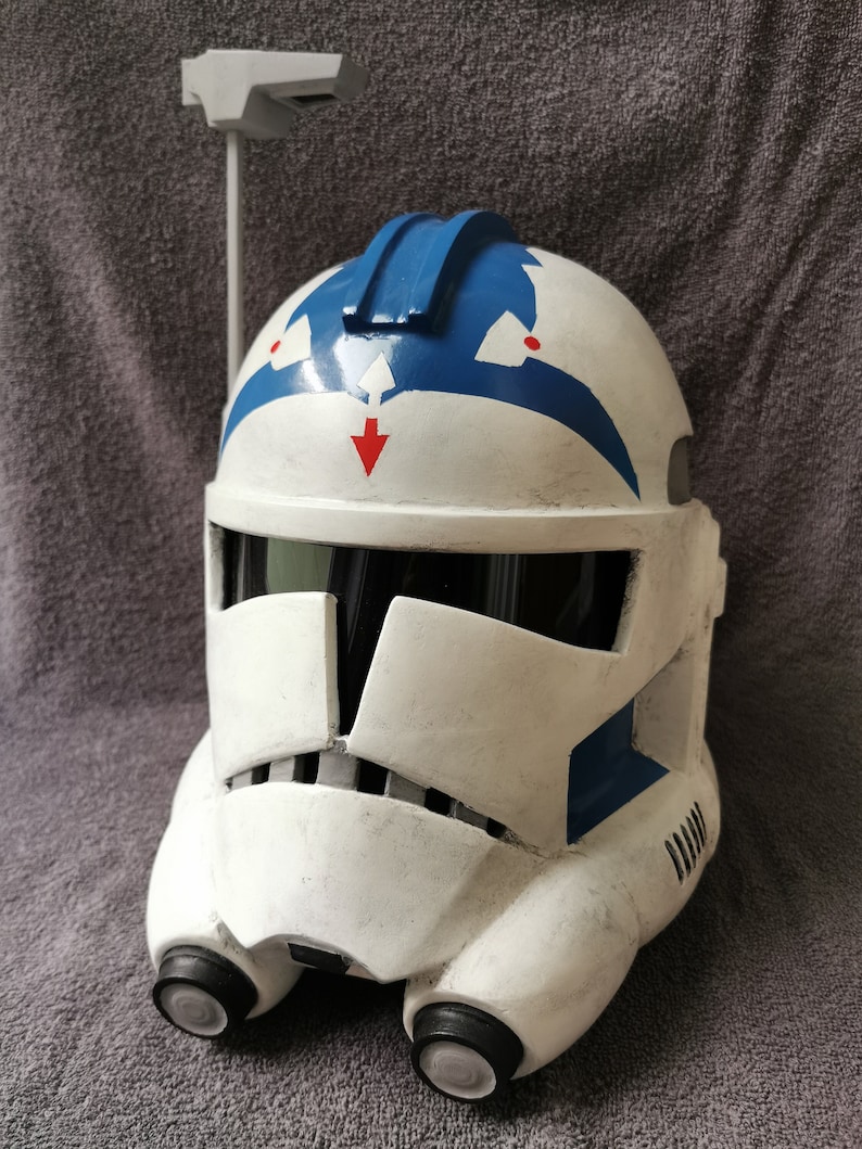 May include: A white and blue Star Wars clone trooper helmet with a red arrow on the front. The helmet has a blue stripe on the top and a black visor.