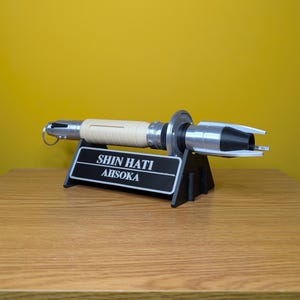 May include: A white and silver lightsaber prop with a black stand. The stand has a black plaque with the text "SHIN HATI AHSOKA" on it.