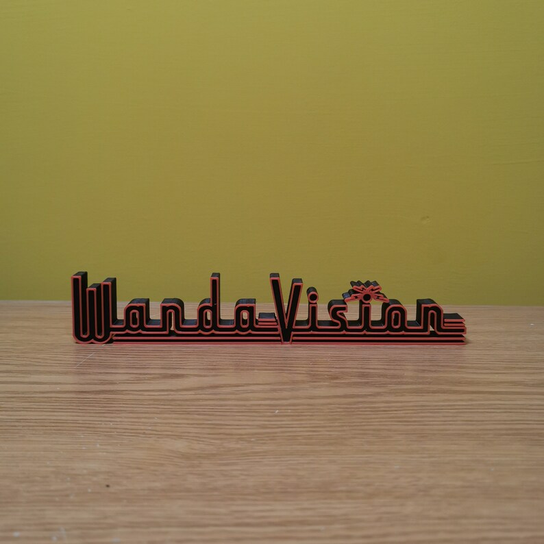 Peut inclure: Panneau imprim&eacute; en 3D avec le texte "WandaVision" dans une police r&eacute;tro. Le signe est noir avec des contours rouges et repose sur une surface en bois. L'arri&egrave;re-plan est de couleur jaune-vert uni.