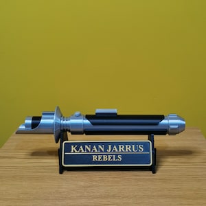 May include: A silver and black lightsaber with a black stand. The stand has a gold plaque that reads "KANAN JARRUS REBELS".