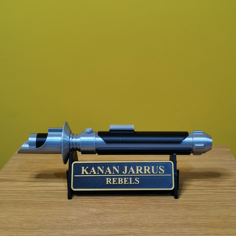 May include: A silver and black lightsaber with a black stand. The stand has a gold plaque that reads "KANAN JARRUS REBELS".