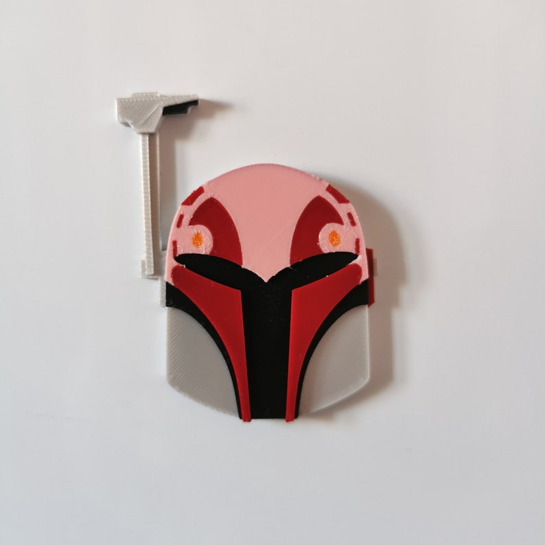May include: A 3D printed Mandalorian helmet with a red and black design. The helmet has a gray base and a pink top with orange accents.
