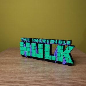K&ouml;nnte beinhalten: Ein 3D-gedrucktes Modell des "The Incredible Hulk"-Logos. Das Logo besteht aus gr&uuml;nem und violettem Kunststoff und hat eine Ziegelstruktur. Das Logo steht auf einer Holzoberfl&auml;che.